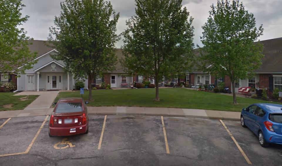 Exterior view of Elliott Place Senior Living showing a single-story brick building with white trim, a covered entrance, green lawn with trees, and a parking lot with two cars, one parked in a handicapped spot.