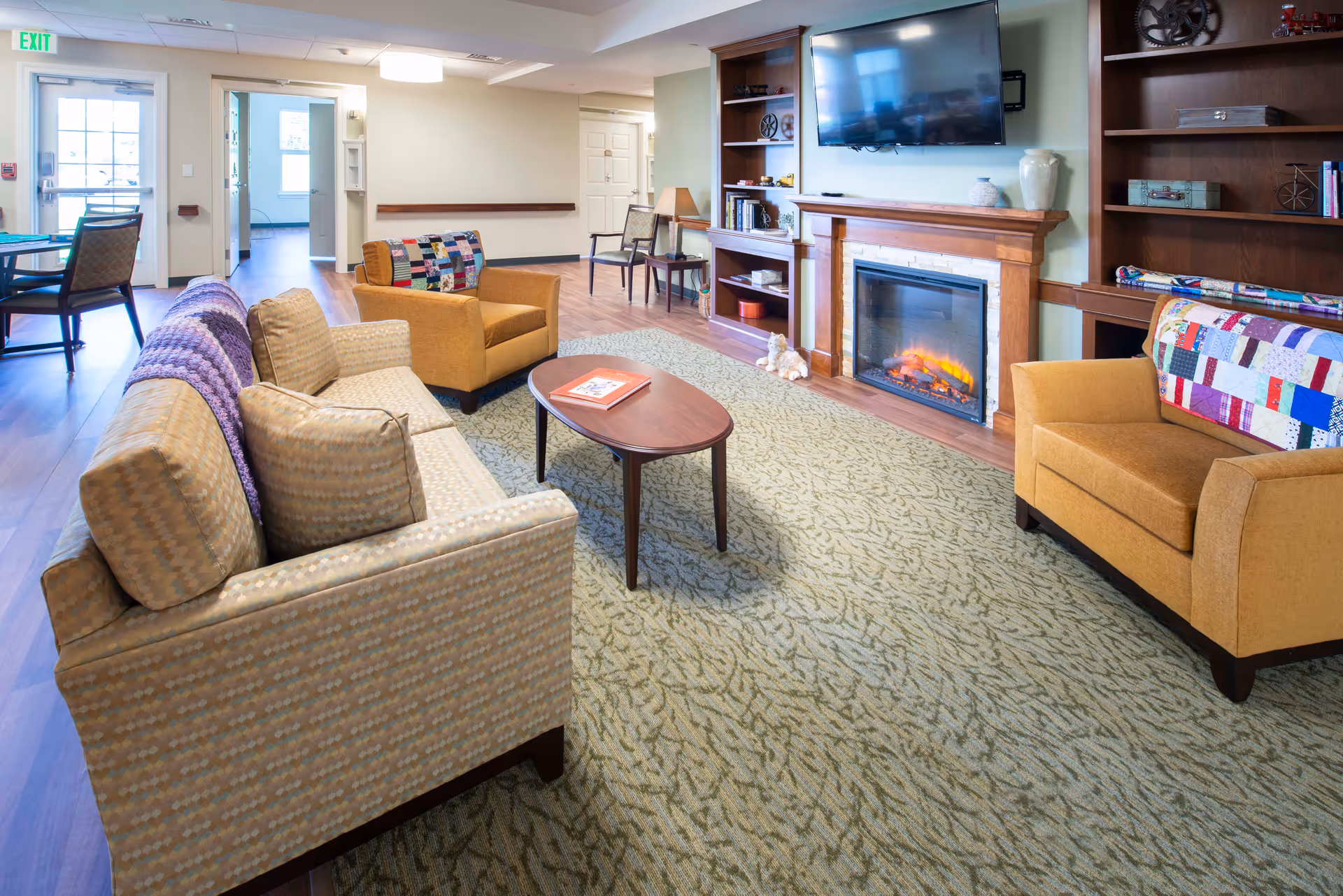 A cozy living room in a senior living facility featuring a patterned sofa and two mustard-colored armchairs with patchwork quilts draped over the backs. A wooden coffee table sits on a green patterned carpet in front of an electric fireplace with a wooden mantel. Above the fireplace is a mounted flat-screen TV. Built-in wooden shelves flank the fireplace, holding decorative items and books. In the background, there is a dining table with chairs near a door marked with an exit sign.