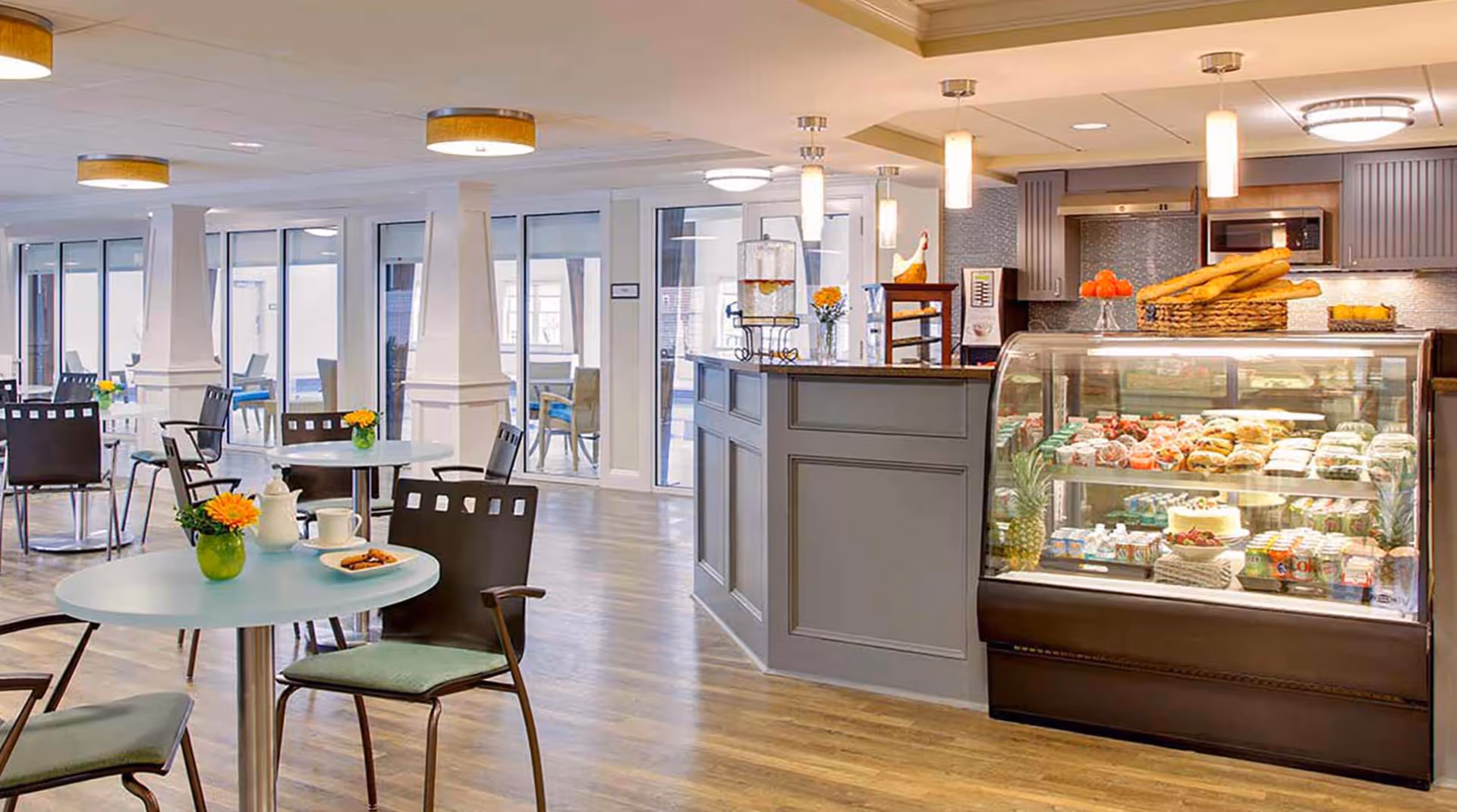 A bright and modern dining area with round tables and chairs, decorated with small vases of yellow flowers. To the right, there is a counter with a glass display case filled with various pastries, cakes, and beverages. The room has large windows and glass doors allowing natural light to enter, and the floor is wooden.