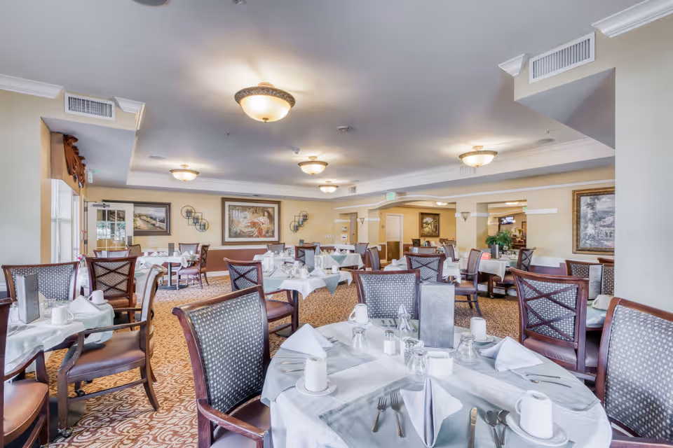 A spacious dining room with multiple tables covered in white and gray tablecloths, each set with napkins, cups, glasses, and silverware. The room features patterned carpet, cushioned wooden chairs, framed artwork on the walls, and ceiling lights providing warm illumination.
