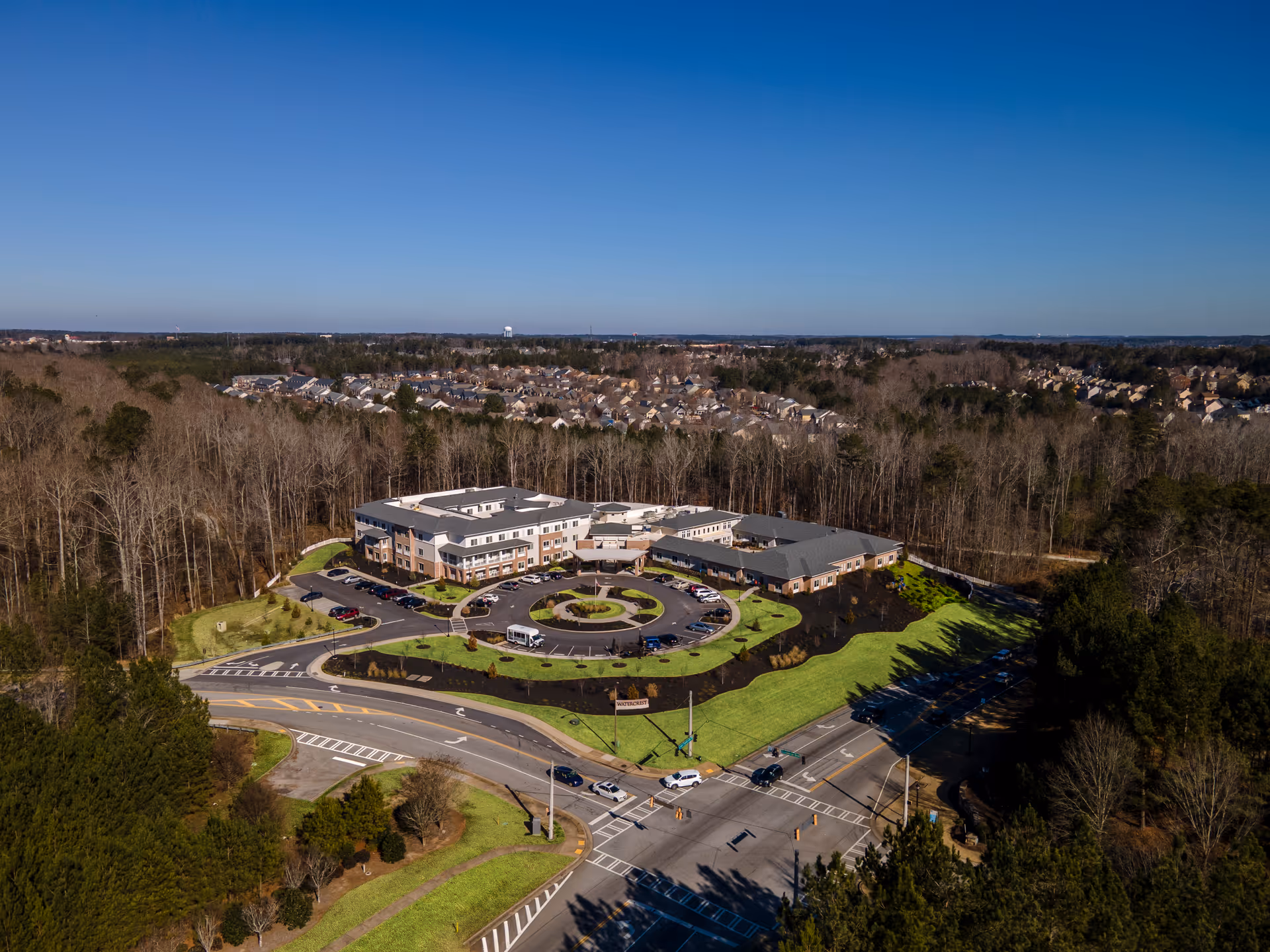 Aerial view of Westhill Newnan Crossing senior living facility surrounded by trees and residential houses in the distance under a clear blue sky. The building has a circular driveway with parked cars and well-maintained landscaping.