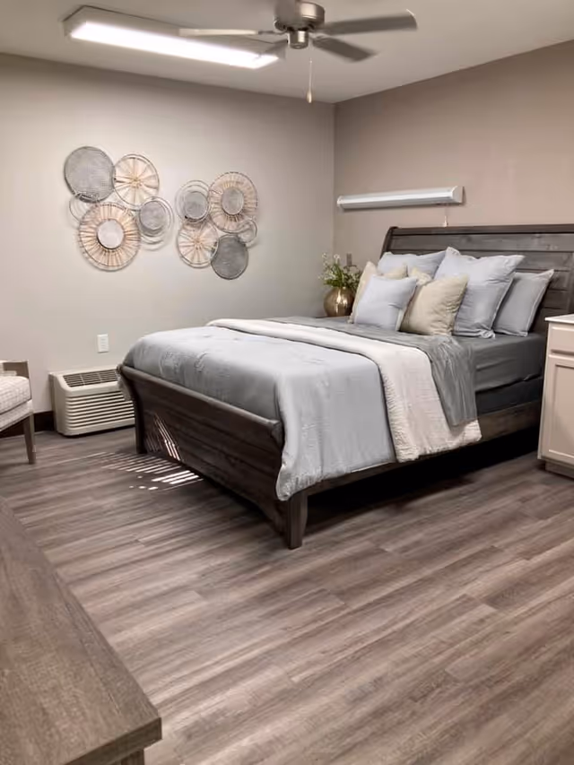 Bed with light blue bedding and multiple pillows in a furnished bedroom with wall art, ceiling fan, and wood-look flooring.