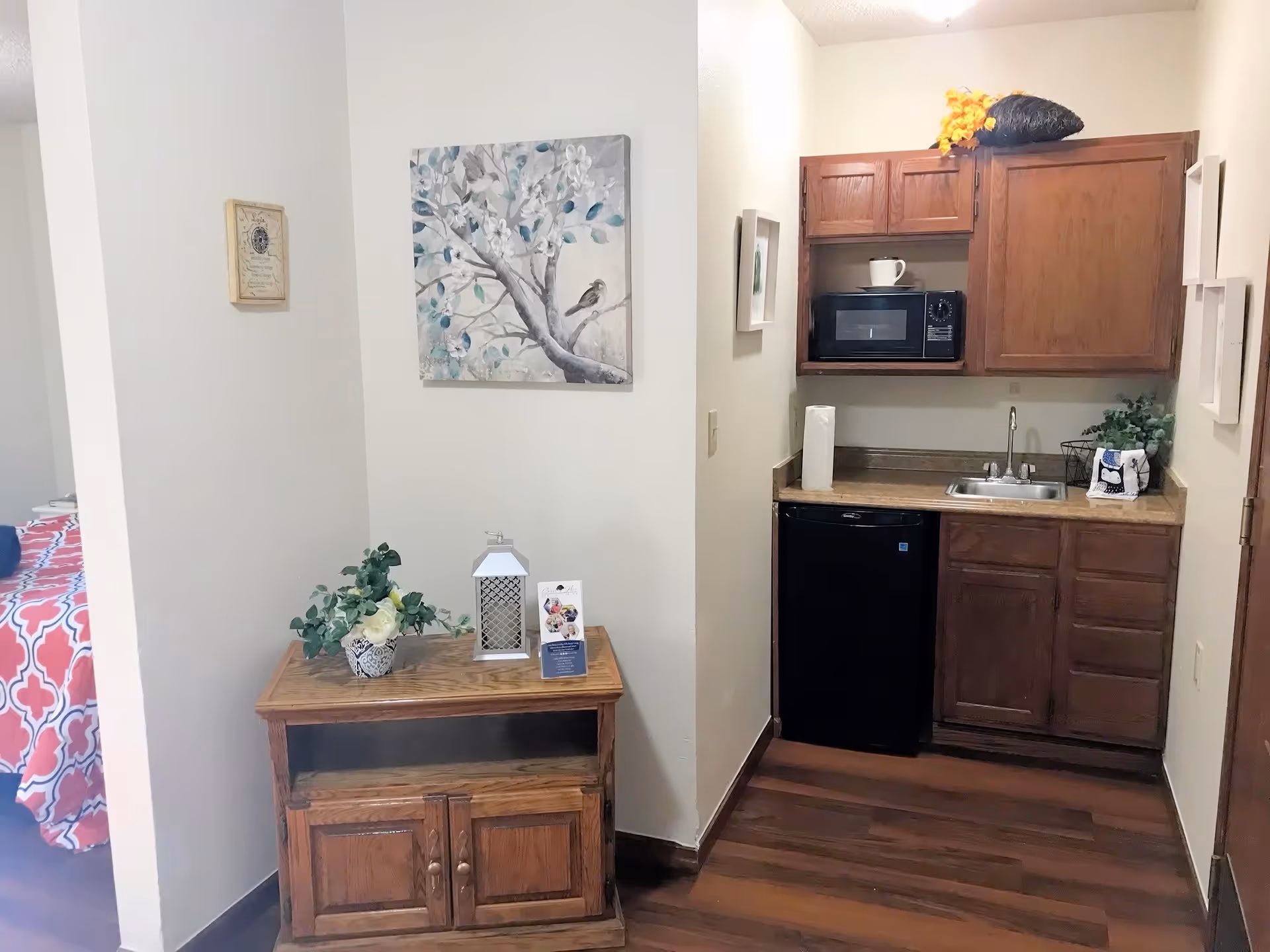 Small kitchenette area with wooden cabinets, a microwave, a mini refrigerator, and a sink. To the left, there is a wooden cabinet with decorative items including a plant, a lantern, and a brochure. A bed with a red and white patterned bedspread is partially visible in an adjacent room.