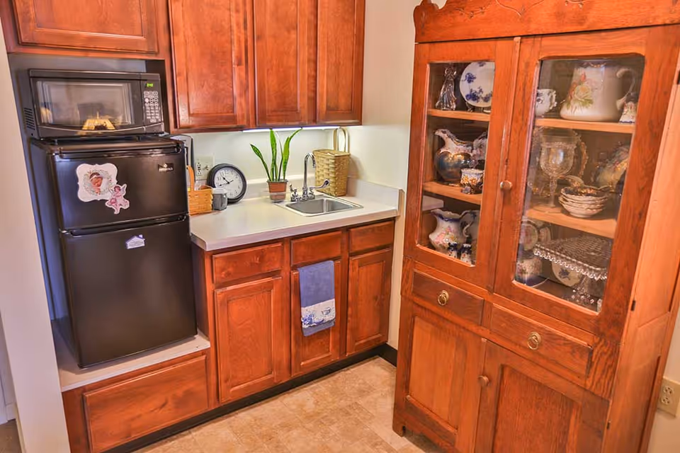 Small kitchen area with wooden cabinets, a black mini refrigerator with a microwave on top, a countertop with a sink, a small plant, a clock, and a towel hanging from the cabinet. To the right, there is a wooden display cabinet with glass doors showcasing decorative dishes and glassware.