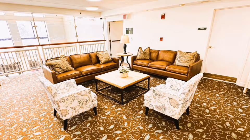 Communal lounge area with two brown leather sofas and two patterned armchairs arranged around a central coffee table on a patterned carpet.