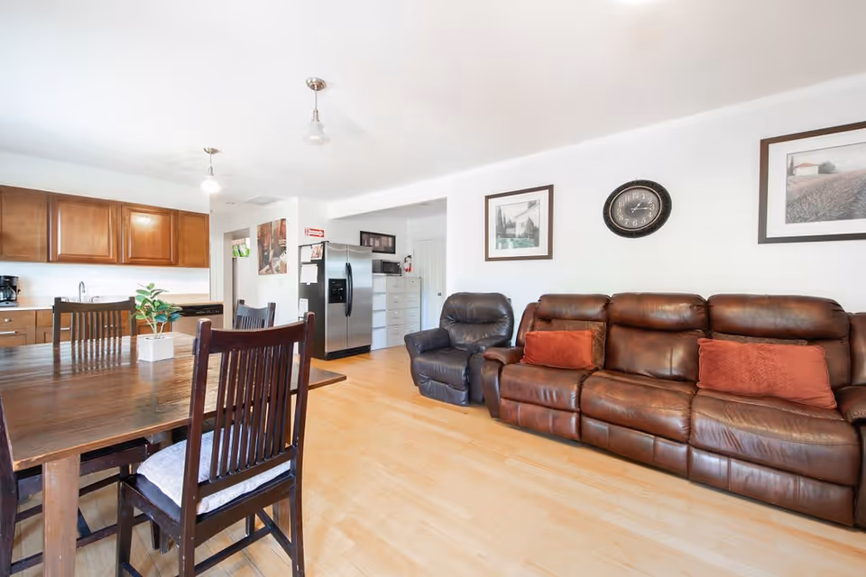 A bright, open living and dining area featuring a wooden dining table with chairs, a brown leather sofa and armchair, a wall clock, framed pictures on the wall, and a kitchen area with wooden cabinets and a stainless steel refrigerator in the background.