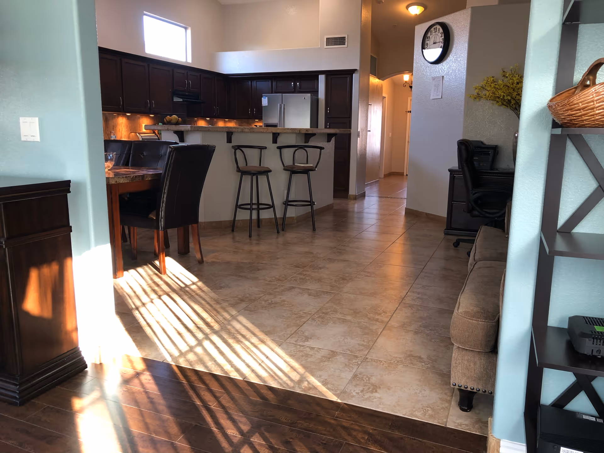 Open-plan interior showing a kitchen with a breakfast bar and stools, adjacent dining table and tiled floor leading to a hallway.