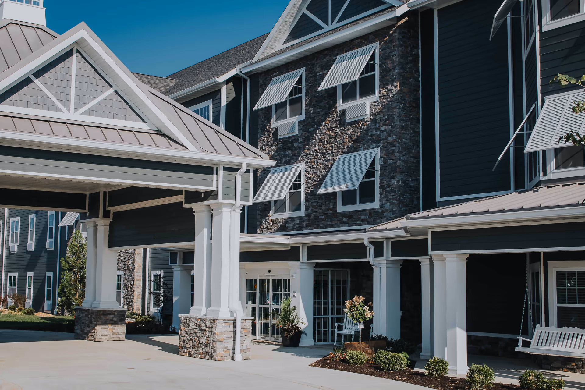 Exterior view of a senior living facility building with stone and dark siding, white columns supporting a covered entrance, and several windows with awnings under a clear blue sky.