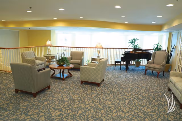 A spacious senior living community lounge area with patterned carpet, several upholstered armchairs arranged around a wooden coffee table with a plant centerpiece, two table lamps on side tables, a grand piano with a bench, and large windows letting in natural light.