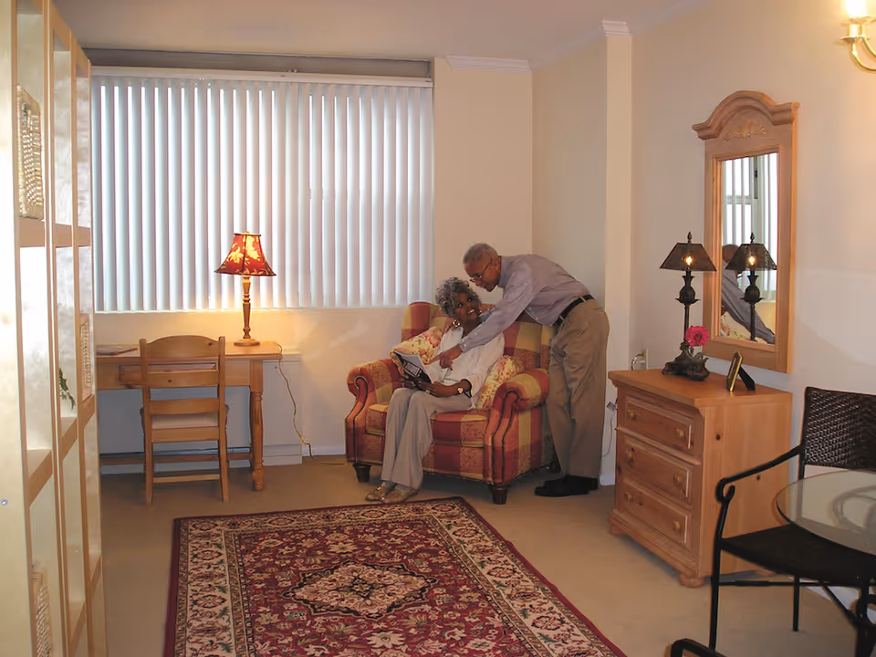 A cozy living room with an elderly couple interacting warmly. The woman is seated on a patterned armchair while the man leans over her. The room features a wooden desk with a chair and a lamp, a wooden dresser with a mirror and two lamps, a glass-top table with a chair, and a decorative rug on the floor. Vertical blinds cover the window.