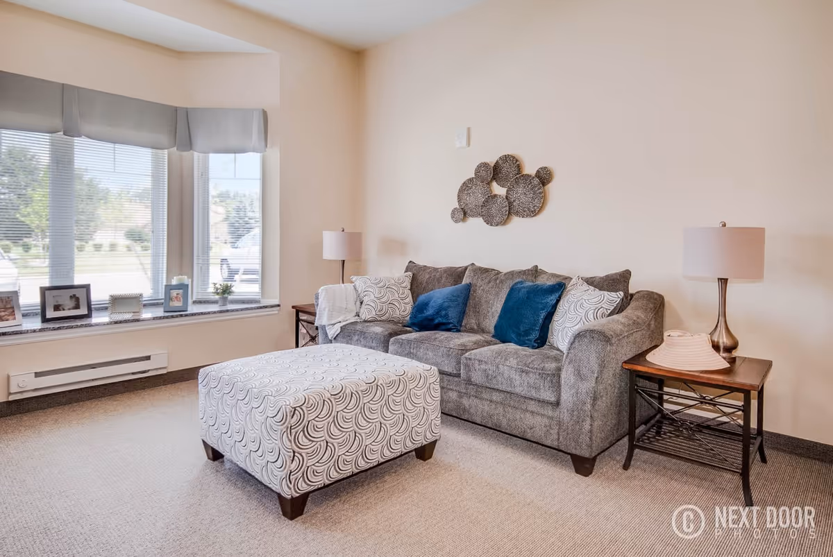 A cozy living room with a gray sofa adorned with blue and patterned pillows, a matching patterned ottoman in front, two side tables each with a lamp, and a large window with blinds and valances letting in natural light. Decorative wall art is hung above the sofa.