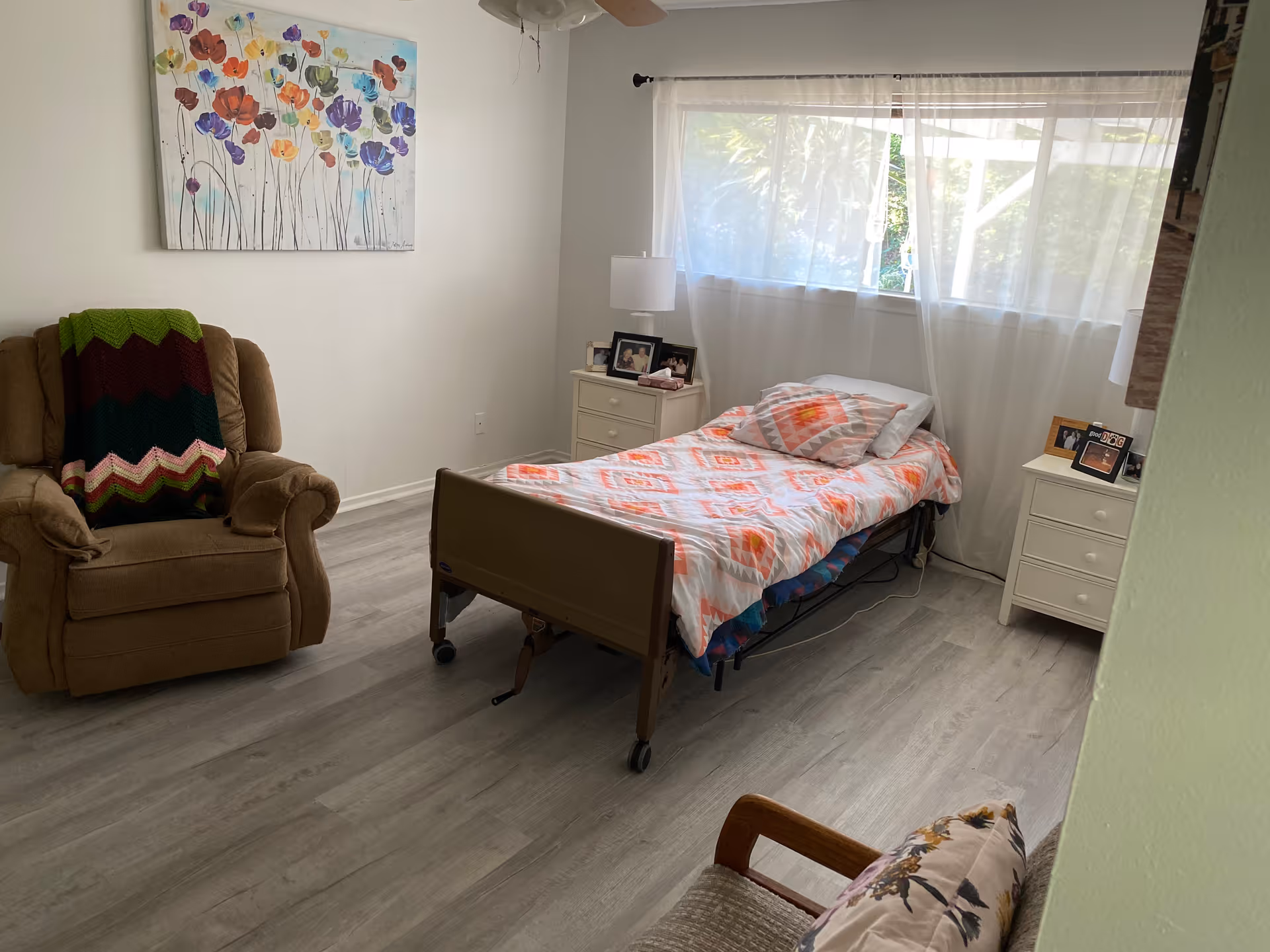 A bright bedroom with a single bed covered in a colorful quilt and pillow, two white nightstands with lamps and framed photos, a brown recliner chair with a multicolored blanket draped over it, a wooden chair with a floral cushion, light gray wood flooring, a large window with sheer white curtains, and a colorful floral painting on the wall.