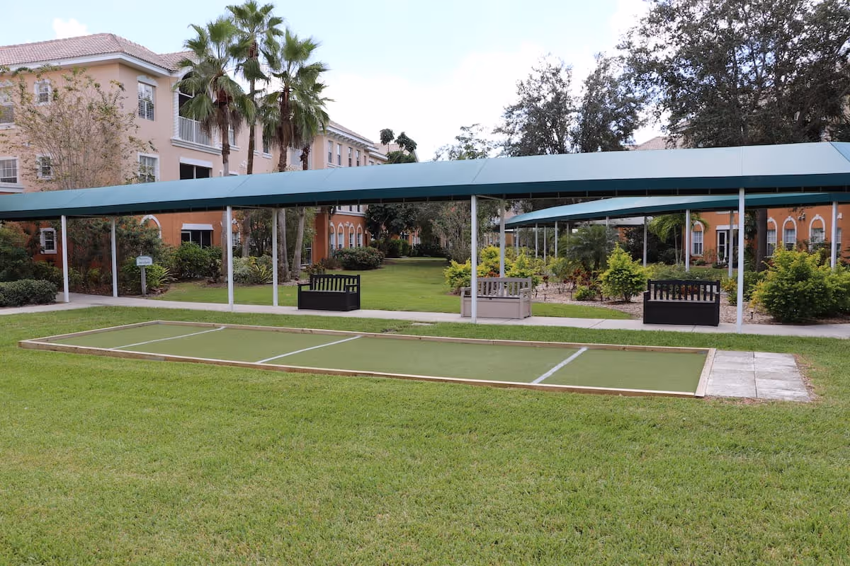 Outdoor area of a senior living facility with a bocce ball court in the foreground, green grass, covered walkways, benches, palm trees, and multi-story buildings in the background.