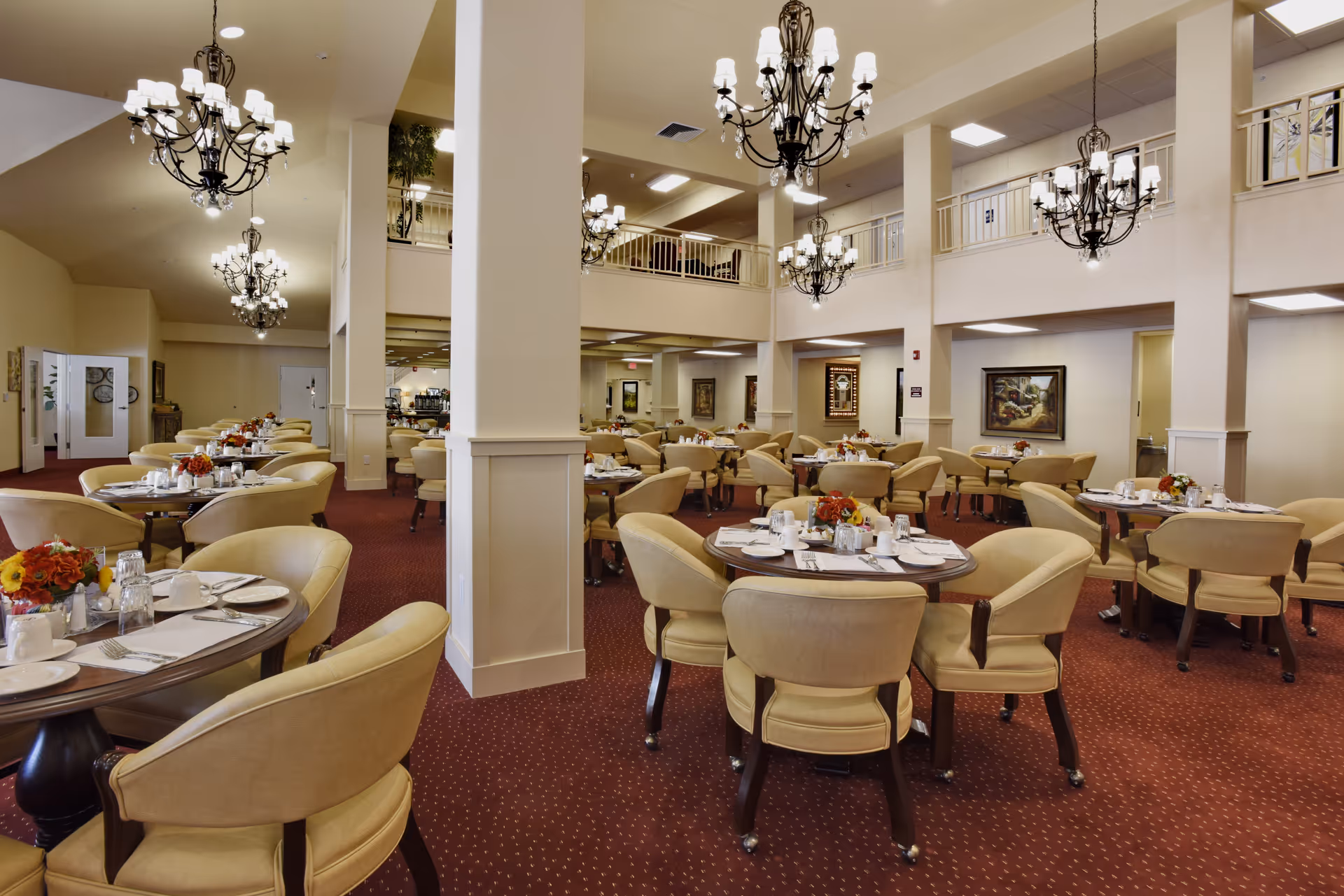 Spacious dining room with round tables set for meals, surrounded by beige upholstered chairs. The room features high ceilings with multiple elegant chandeliers, red carpet flooring, and light-colored walls with framed artwork. There is a second-floor balcony overlooking the dining area.