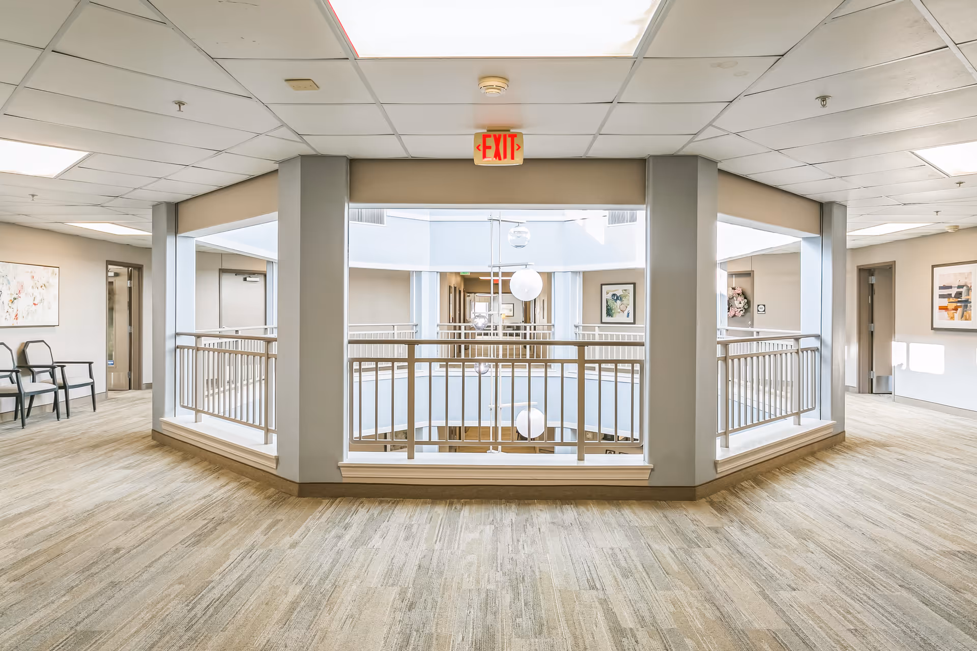 Interior view of a senior living facility hallway with beige walls and carpeted floors. The hallway surrounds an open atrium with railings and hanging spherical light fixtures. There are chairs and framed artwork on the walls, and an illuminated exit sign is visible above the atrium opening.