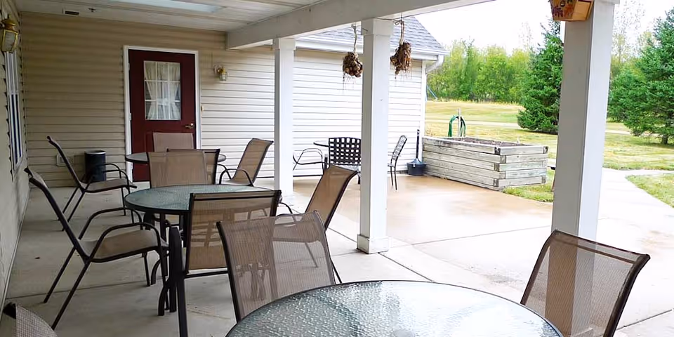 Covered outdoor patio with glass-top tables and mesh chairs facing a lawn and raised garden beds.