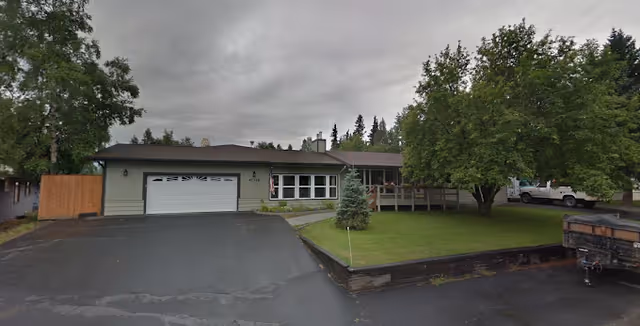 Front exterior of a single-story assisted living building with an attached garage, driveway, lawn, and trees under a cloudy sky.