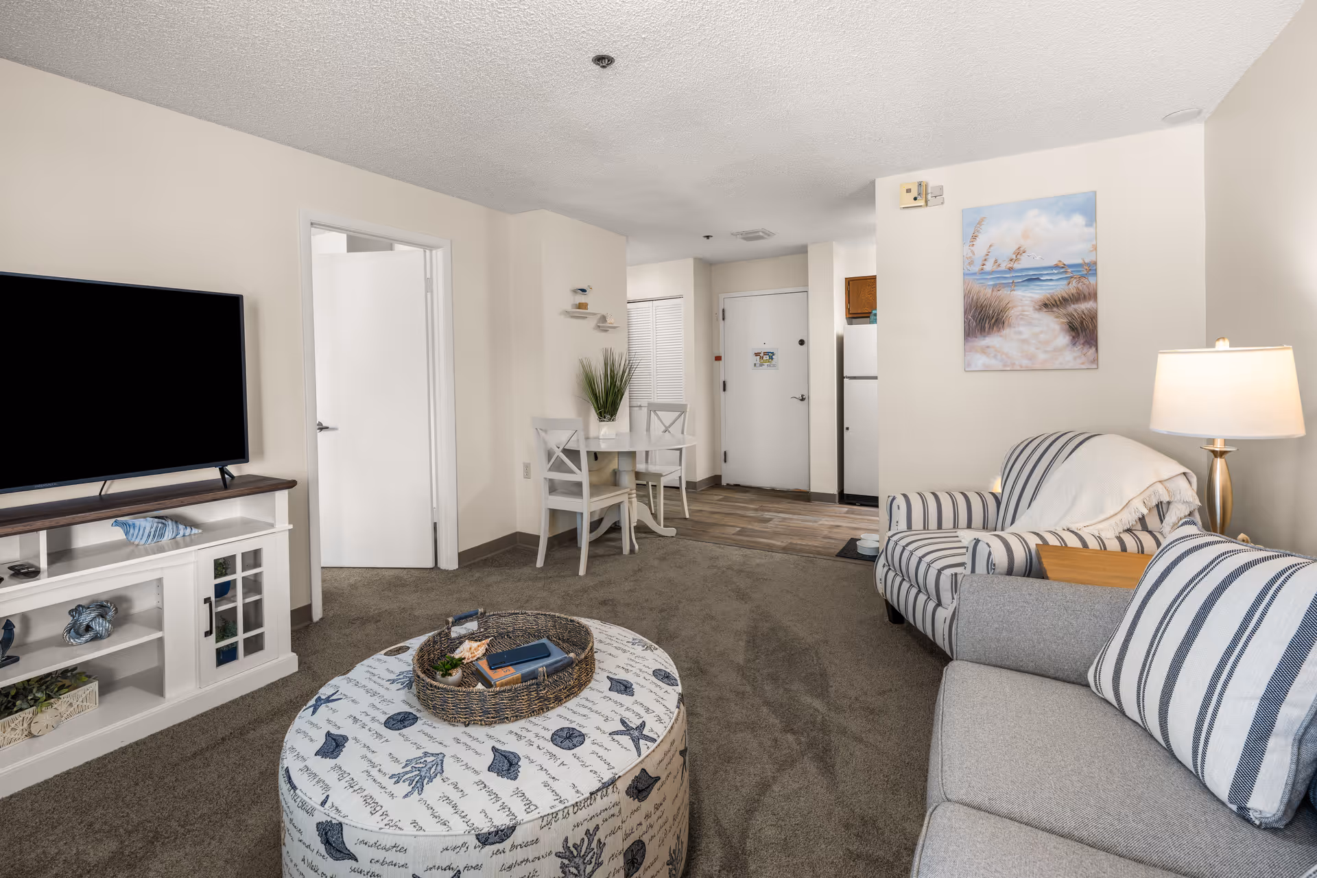 A cozy living room in a senior living facility featuring a gray sofa with striped pillows, a matching striped armchair, a round ottoman with a nautical-themed cover, a white TV stand with decorative items, a flat-screen TV, a small dining table with two chairs, a floor lamp, and a painting of a beach scene on the wall. The entrance door and part of the kitchen with a refrigerator are visible in the background.