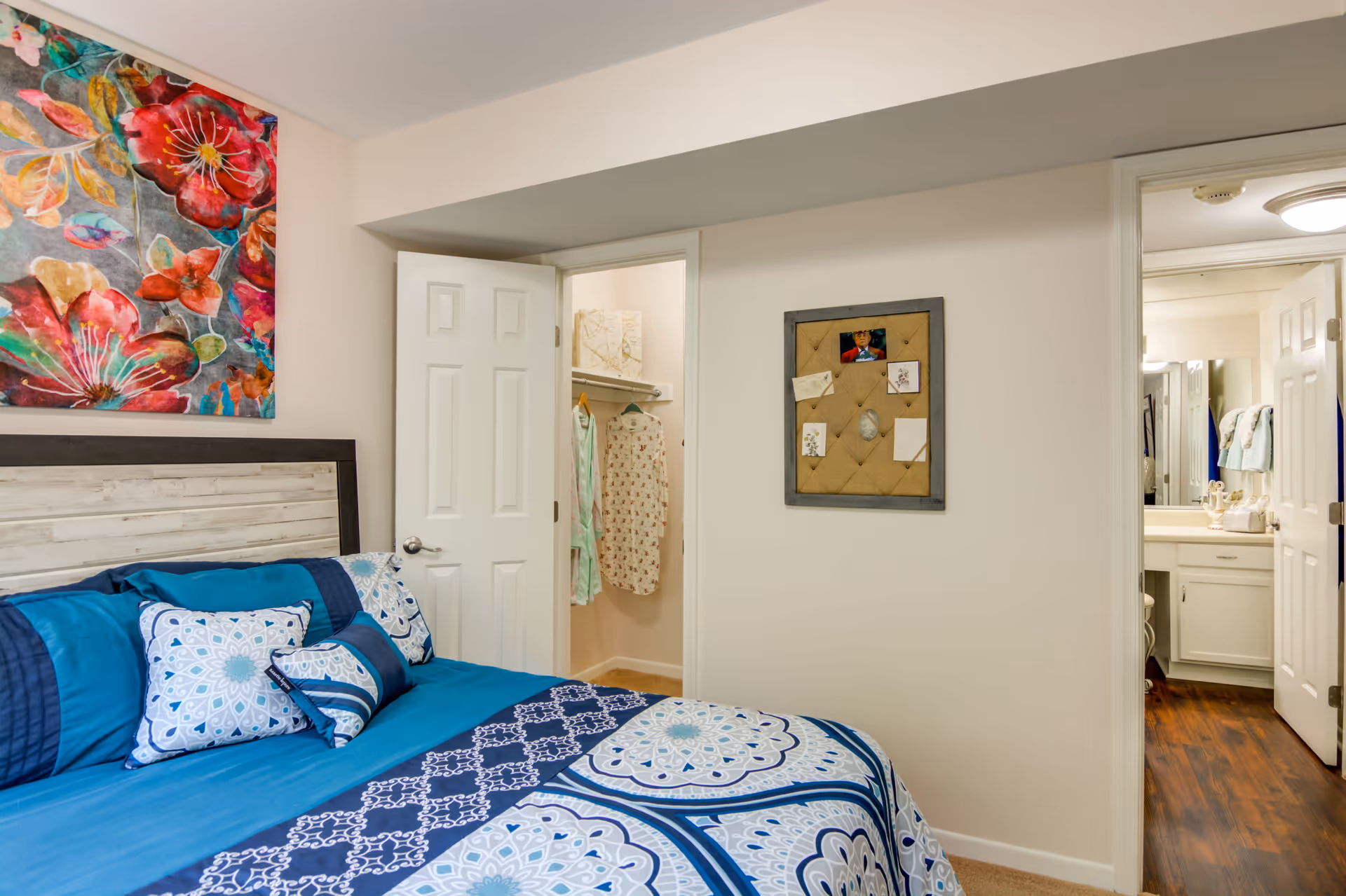 A bedroom with a bed made up with blue and white patterned bedding and multiple pillows. Above the bed is a colorful floral painting. To the right, there is an open door leading to a walk-in closet with clothes hanging inside. Another open door reveals a bathroom with a sink and mirror. The walls are light-colored and the floor in the bathroom area is wooden.