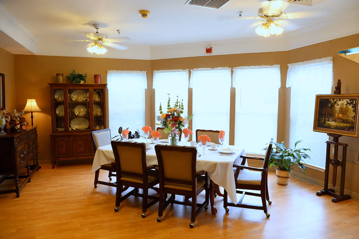 A well-lit dining room with a rectangular table covered with a white tablecloth, set with plates, glasses, and napkins folded in a fan shape. Six chairs surround the table. The room has wooden flooring, large windows with white blinds, two ceiling fans with lights, a wooden cabinet displaying plates, a sideboard with floral arrangements, a potted plant, and a framed painting on an easel.