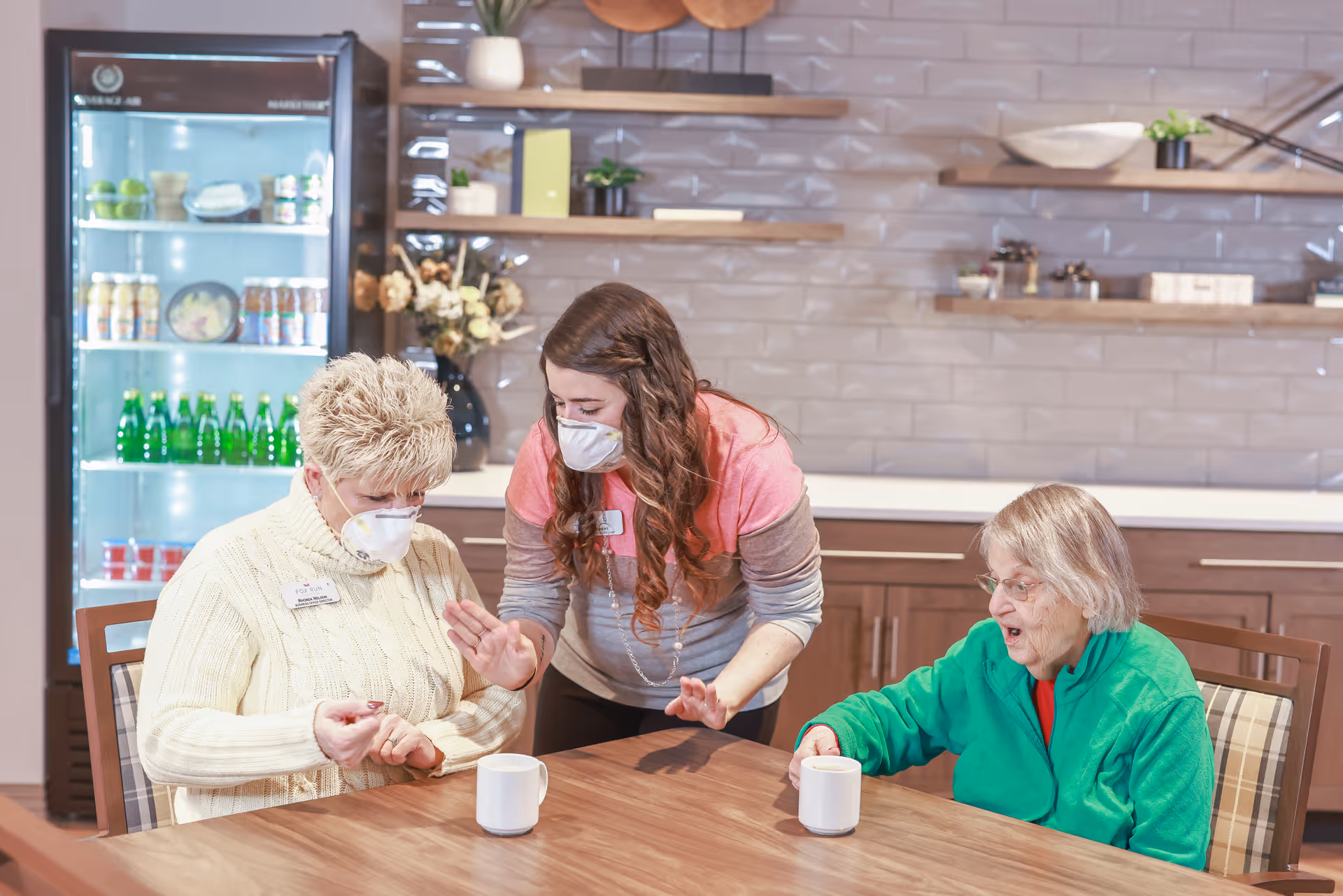 A masked caregiver helps two masked elderly women seated at a table with mugs in a kitchen-style dining area.