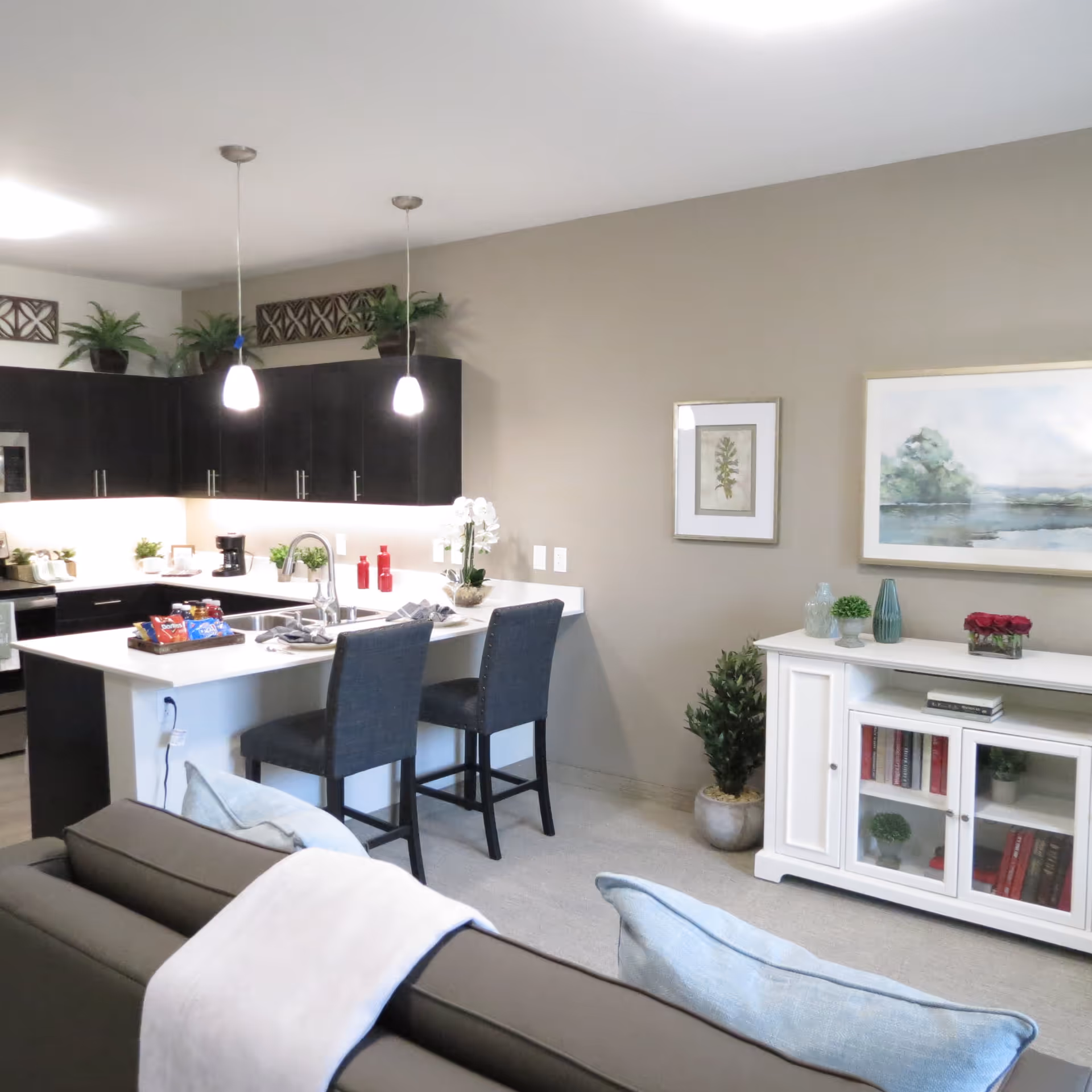Open-plan living area and kitchen with a white island, two bar stools, dark cabinets and a sofa in the foreground.