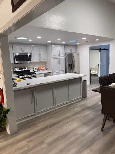 Modern kitchen with white cabinets, stainless steel appliances including a microwave, stove, and refrigerator. A long white countertop island is in the foreground with a few items on it. The kitchen opens into a dining area with a dark dining table and chairs. A sliding barn door leads to another room.