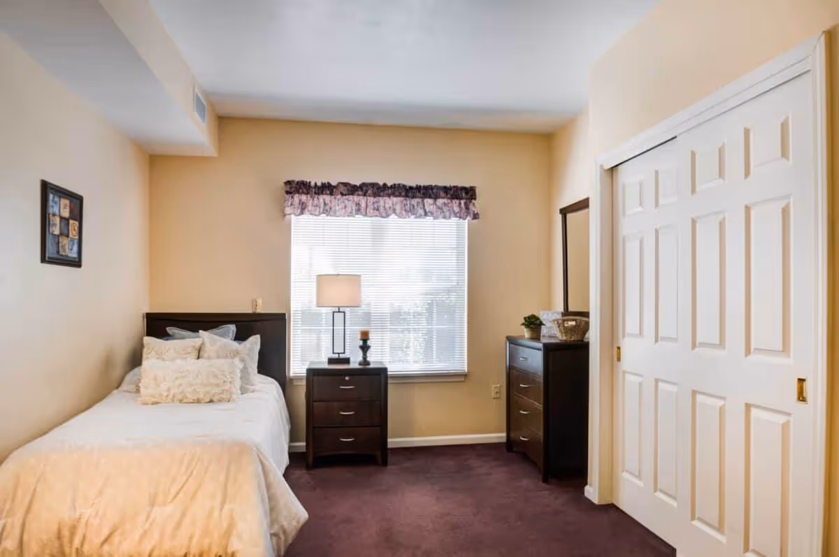 A small bedroom with a single bed on the left side, adorned with multiple pillows and a light-colored blanket. Next to the bed is a dark wooden nightstand with a lamp and a candle. A window with blinds and a floral valance is centered on the back wall. To the right, there is a dark wooden dresser with a mirror above it and a small plant and basket on top. The room has beige walls and a dark carpeted floor. A white sliding closet door is visible on the right side.