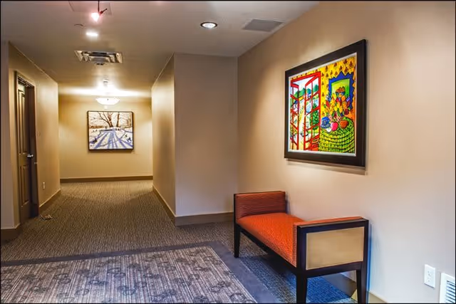 Interior hallway of a senior living facility with beige walls and carpeted floors. A red cushioned bench is placed against the right wall beneath a colorful framed painting. At the end of the hallway, there is another framed artwork depicting a winter scene with trees and shadows on snow. The hallway is softly lit with ceiling lights.