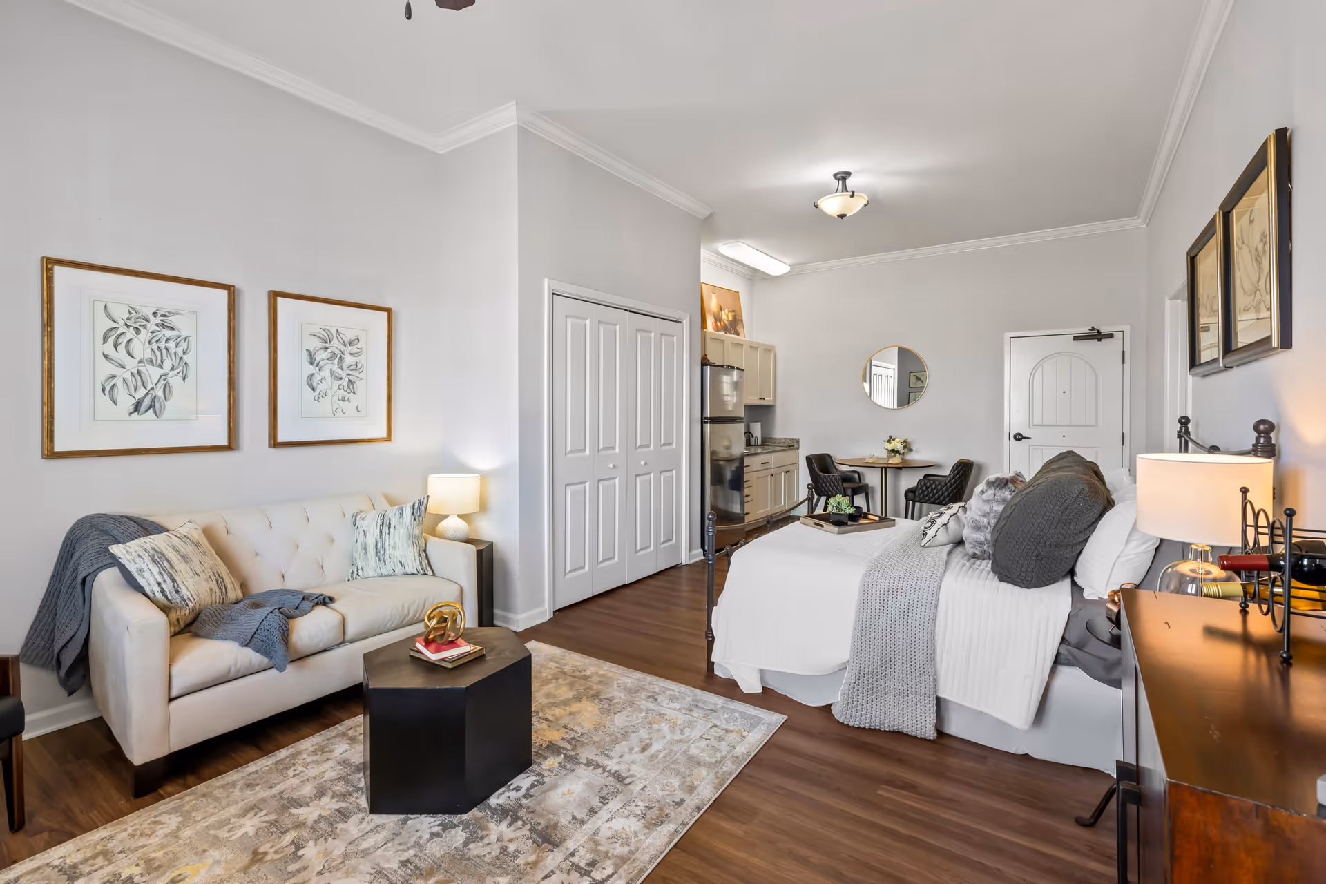 A cozy studio apartment featuring a bed with gray and white bedding, a beige tufted sofa with pillows and a throw blanket, a small black coffee table, a wooden dresser with a lamp, and a small dining area with two chairs and a round table. The room has light gray walls, wood flooring, and framed botanical artwork on the walls.