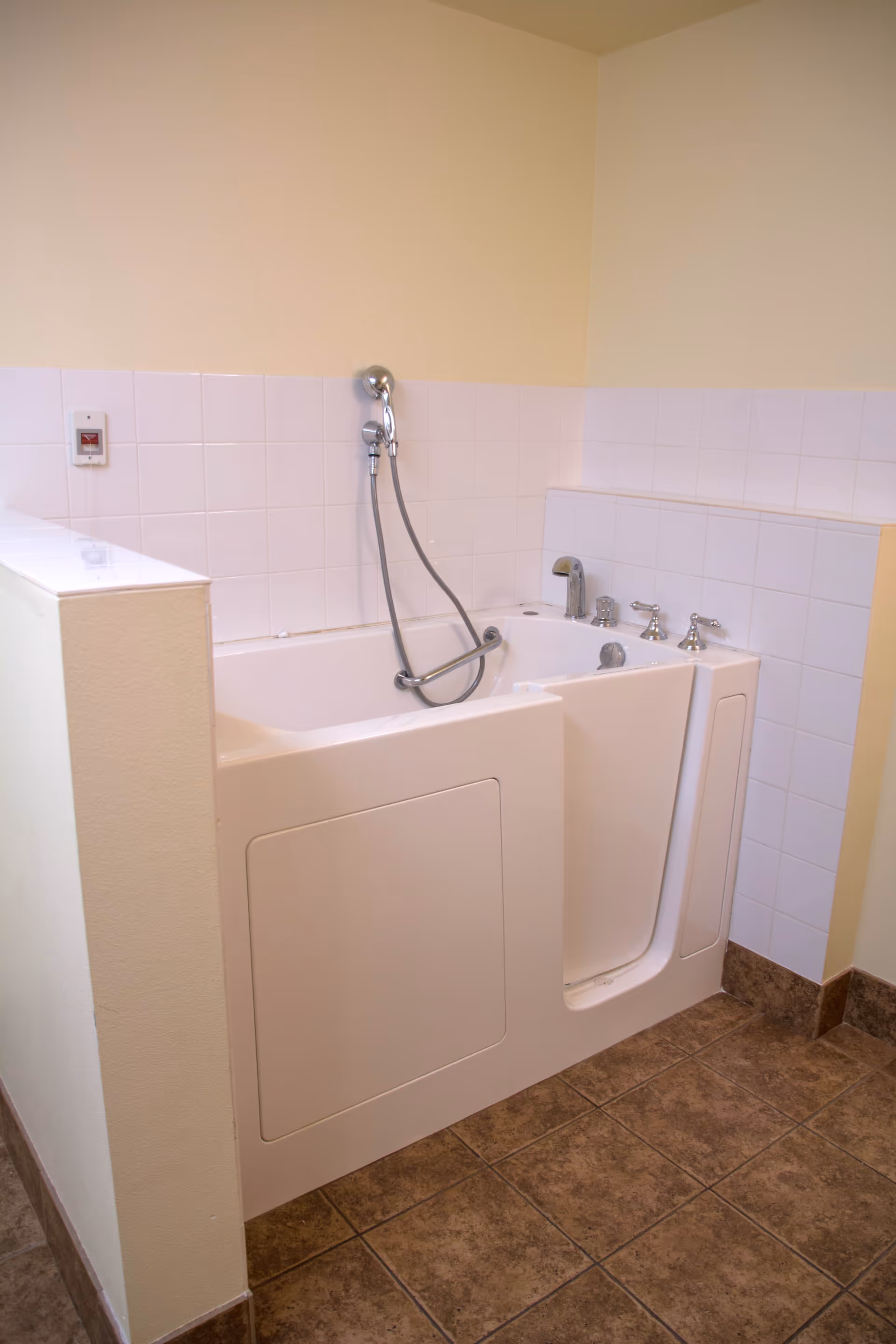 A walk-in bathtub with a door for easy access, equipped with a handheld showerhead and multiple faucet controls, set against a tiled wall in a bathroom with beige walls and brown tiled floor.
