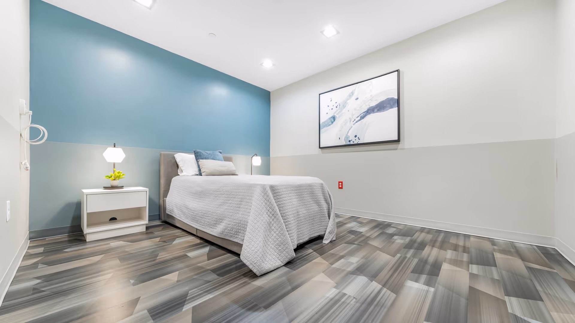 Minimal modern single-occupancy bedroom with a bed, nightstand, lamps, blue accent wall and abstract wall art.