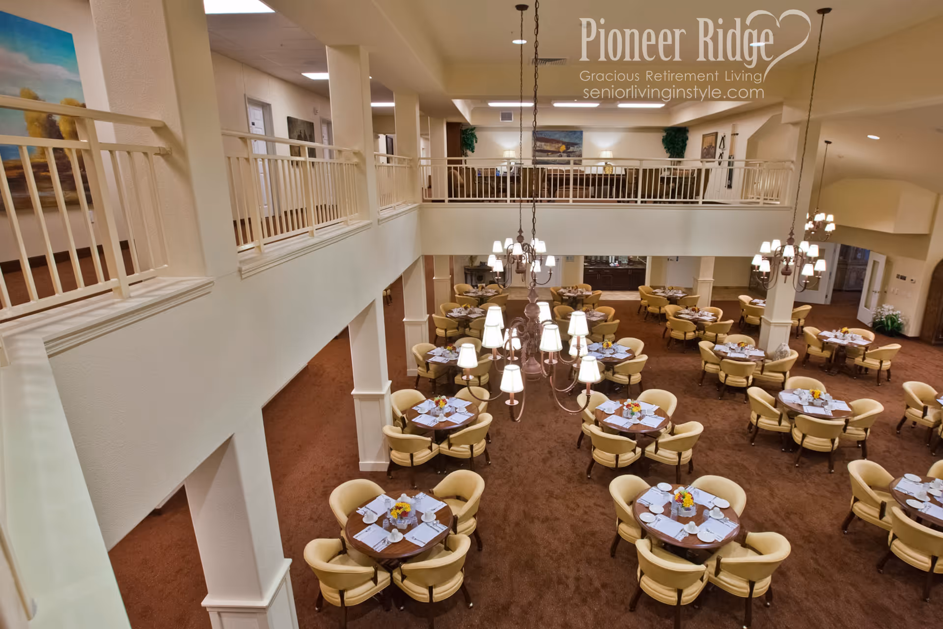 A spacious dining room in a retirement living facility with multiple round tables, each surrounded by comfortable beige chairs. The tables are set with placemats, napkins, cups, and small flower arrangements. The room features warm lighting from chandeliers hanging from the ceiling, and there is a second-floor balcony overlooking the dining area. The walls are decorated with paintings and plants, creating a cozy and inviting atmosphere.
