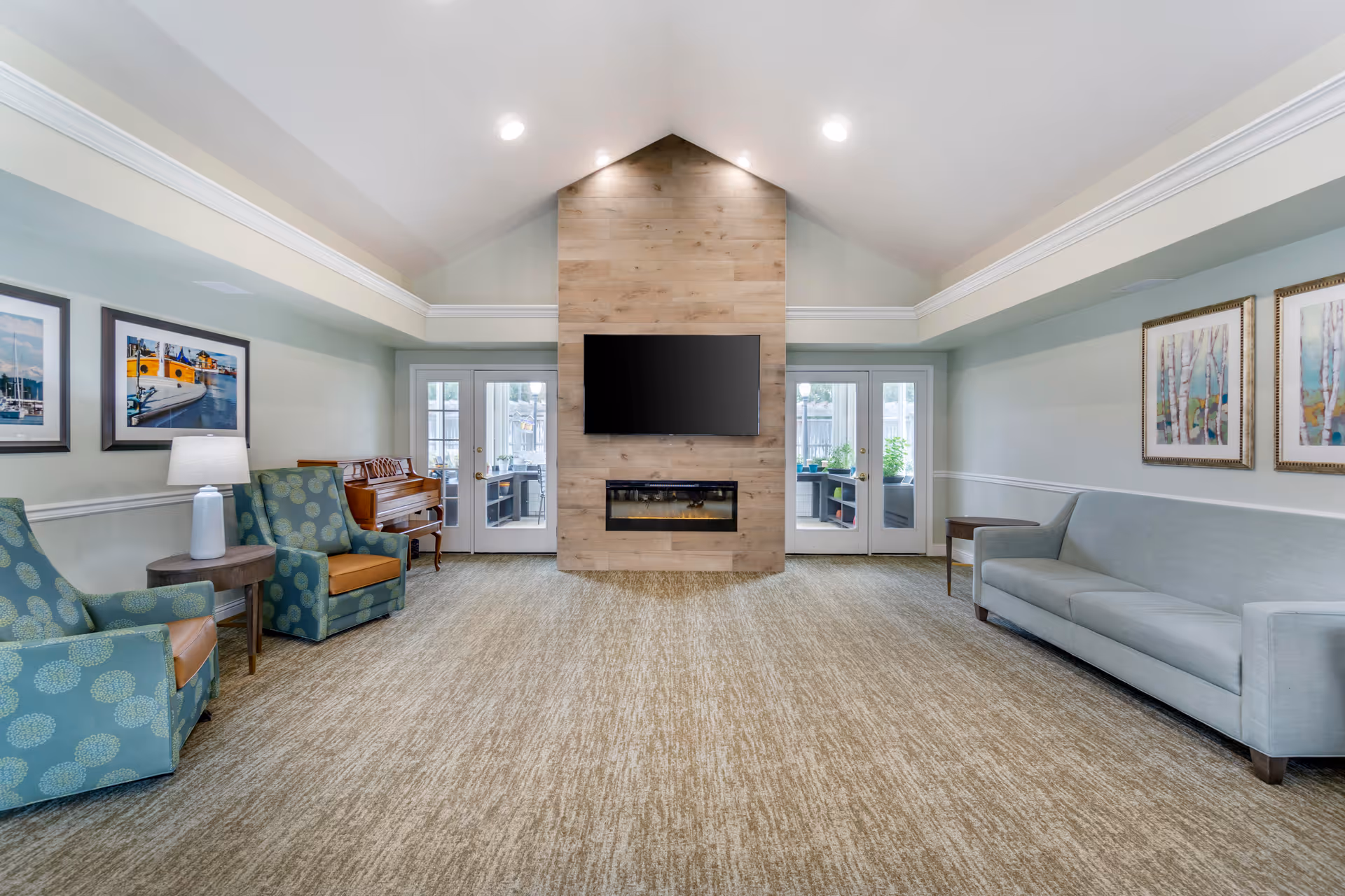 A spacious living room with a vaulted ceiling featuring recessed lighting. The room has a central wooden accent wall with a mounted flat-screen TV above a modern electric fireplace. On the left side, there are two patterned armchairs with a small round table and a lamp between them, and a piano in the corner. On the right side, there is a light gray sofa with two framed artworks above it and a small side table. French doors with glass panes lead to an outdoor area.
