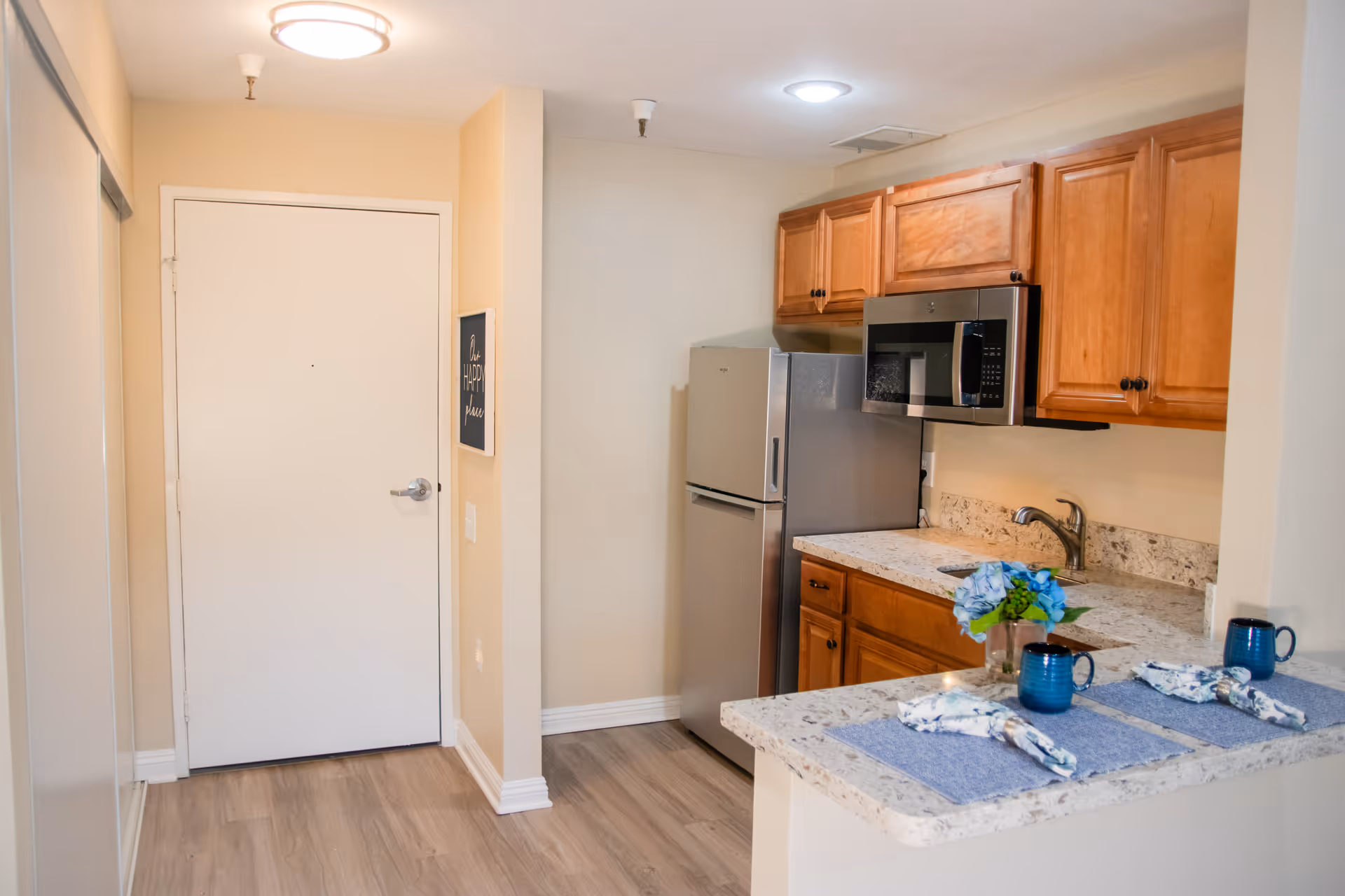 A small kitchen area with wooden cabinets, a stainless steel refrigerator, and a microwave. There is a granite countertop with a sink and a small dining bar set with two blue mugs, napkins, and a vase with blue flowers. The floor is light wood, and there is a white door and a wall sign that says 'Our Happy Place'.