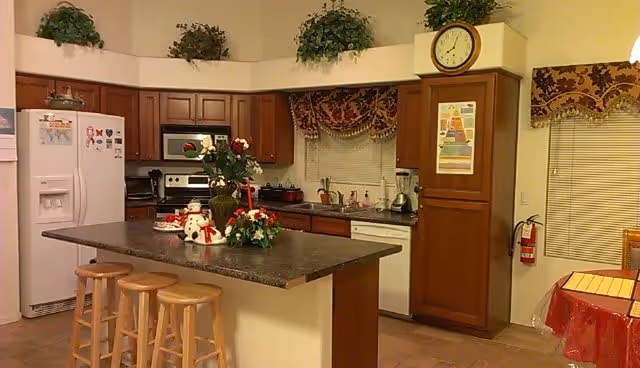 Kitchen with a central island and three wooden stools, wooden cabinets, refrigerator, stove, sink, and decorative plants and holiday decorations.