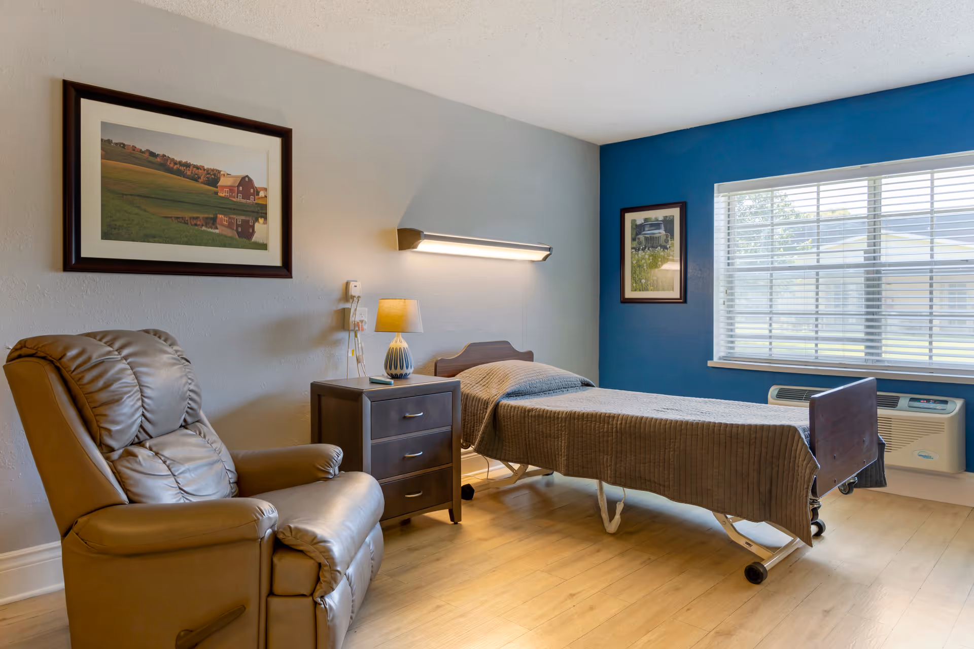 A simple, clean room with a single hospital-style bed covered with a brown blanket, a brown leather recliner chair, a dark wooden nightstand with a lamp, and two framed pictures on the walls. There is a large window with blinds letting in natural light and a wall-mounted light above the bed.