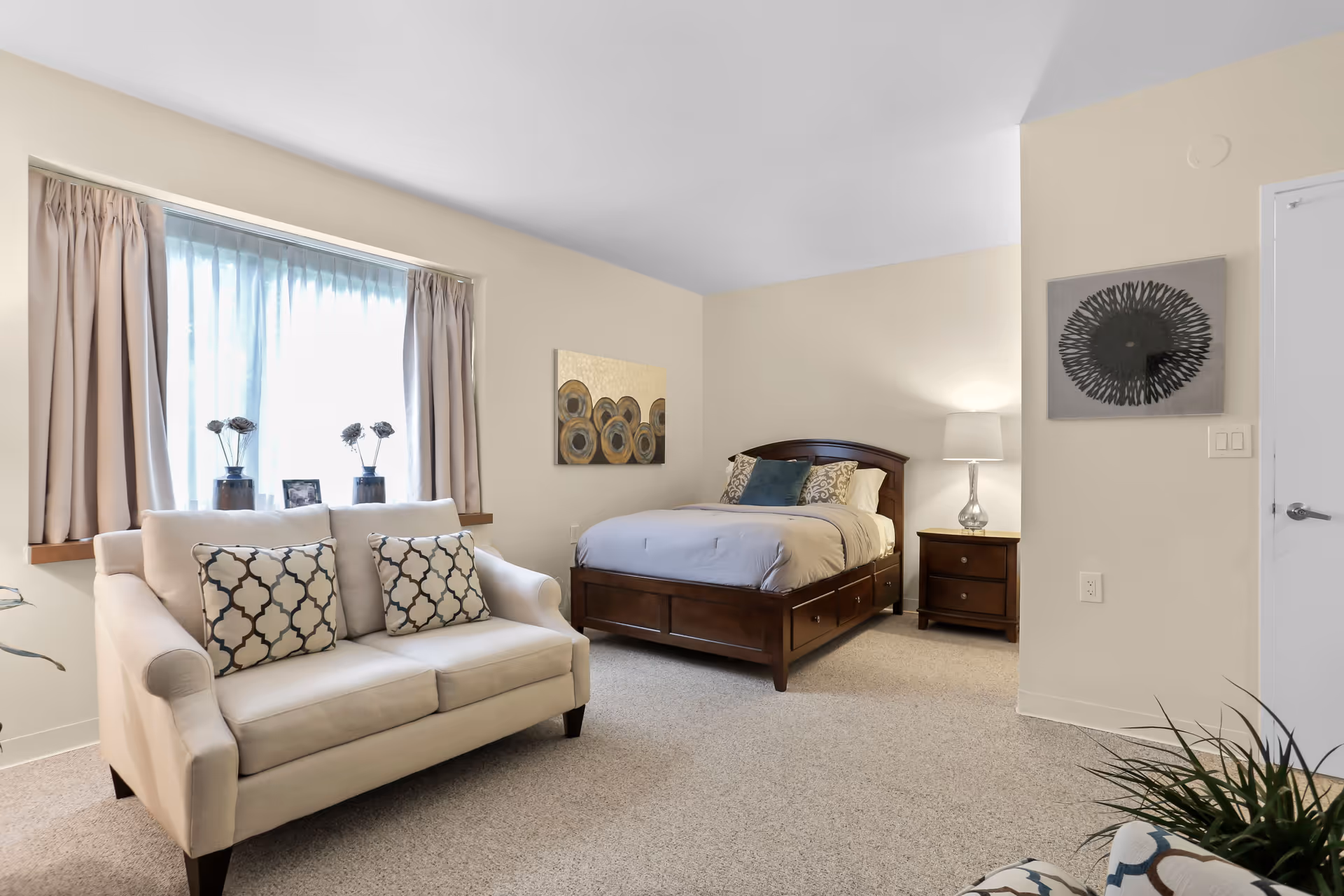 A cozy bedroom with a wooden bed featuring storage drawers underneath, a beige loveseat with patterned pillows in front of a window with curtains, a wooden nightstand with a lamp, and two pieces of wall art. The room has light-colored walls and carpeted flooring.