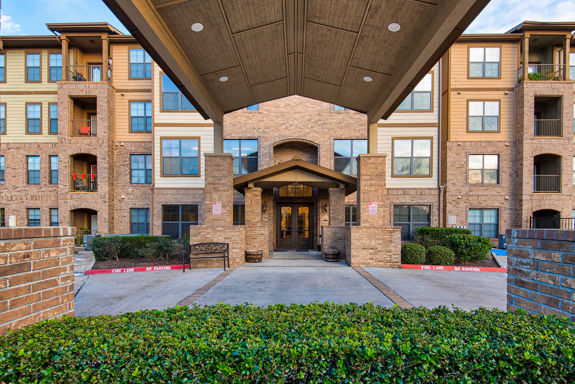 Front entrance of a multi-story residential building with brick and beige siding exterior. The entrance is covered by a large canopy supported by brick pillars. There are bushes and a bench near the entrance, and the driveway has red curbs marked with 'FIRE LANE NO PARKING'.
