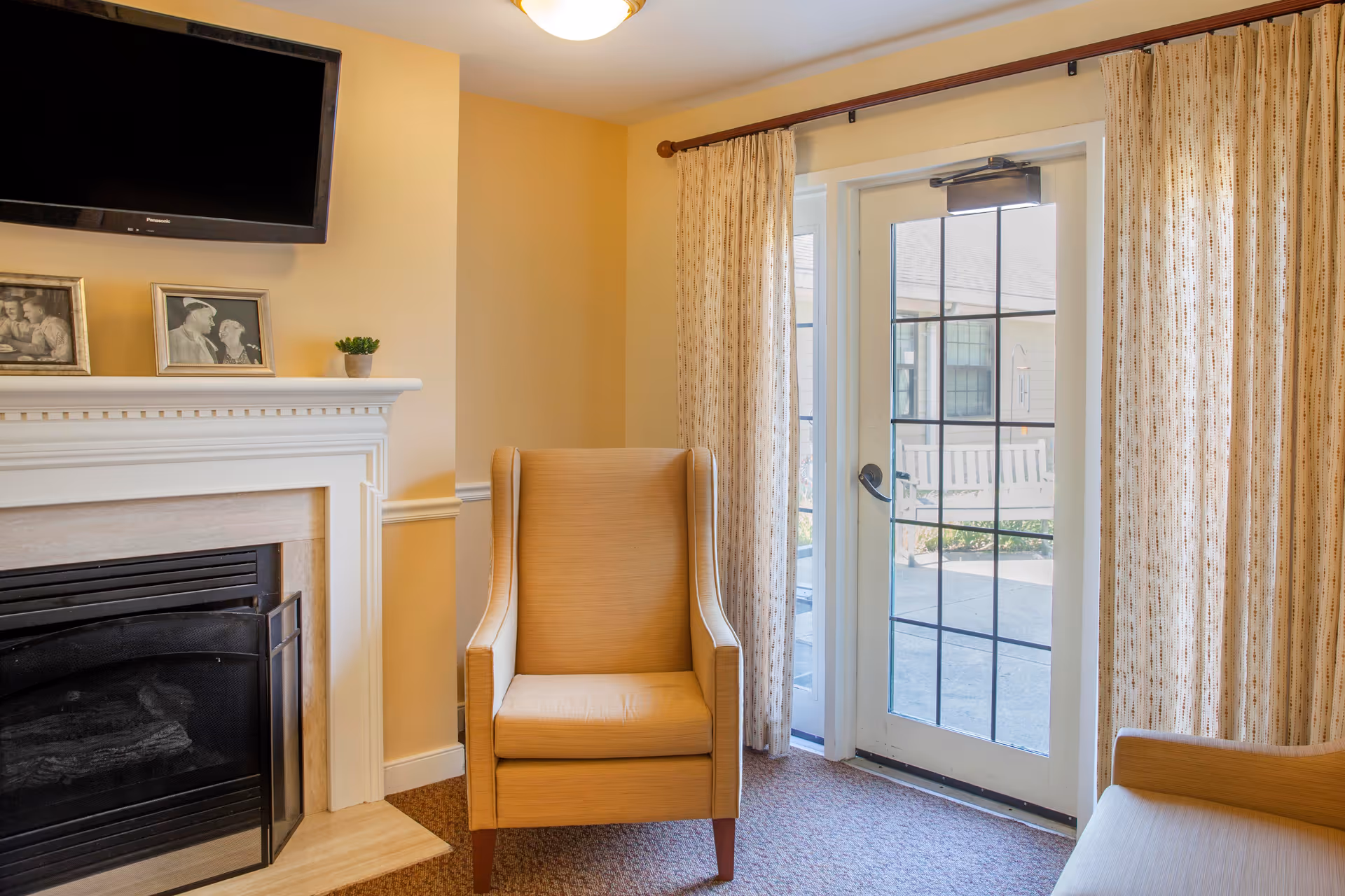A cozy living room area with a beige armchair and a fireplace with a white mantel. Above the mantel is a mounted flat-screen TV and two framed black-and-white photos. To the right, there is a glass door with a grid pattern leading outside, flanked by beige patterned curtains.