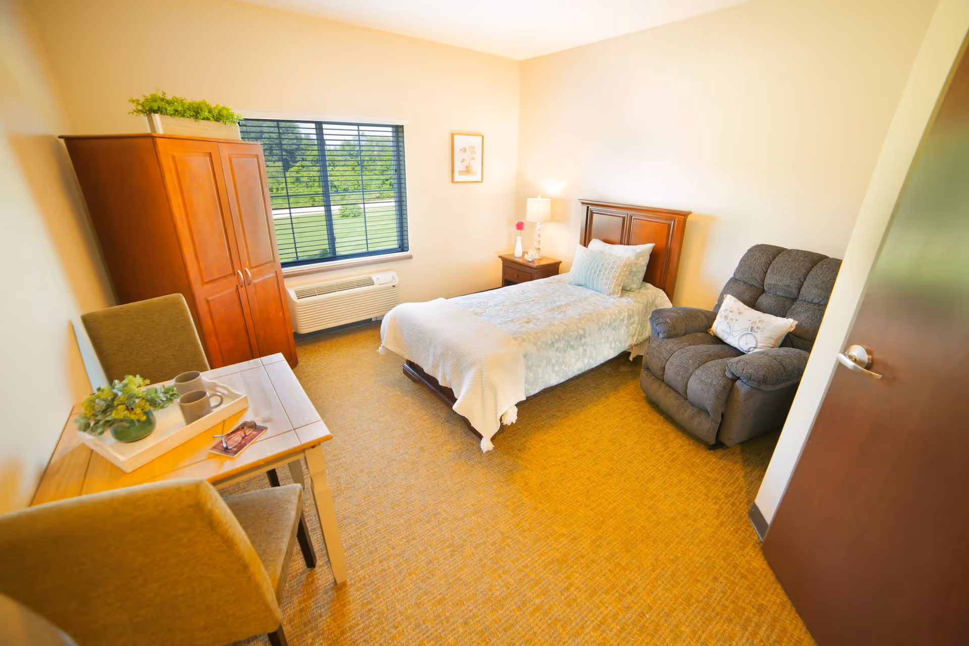 A cozy bedroom with a single bed covered in light blue and white bedding, a wooden nightstand with a lamp and a flower vase, a gray recliner chair with a decorative pillow, a wooden wardrobe, a small table with two chairs, and a window with blinds showing greenery outside.