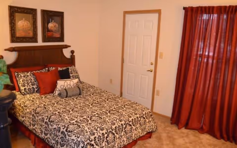 A bedroom with a bed featuring a black and white patterned bedspread and multiple decorative pillows in black, white, and red. The bed has a wooden headboard. There are two framed pictures hanging above the bed. To the right, there is a white door with a brass handle and a window covered by red curtains. The room has beige carpet and light-colored walls.