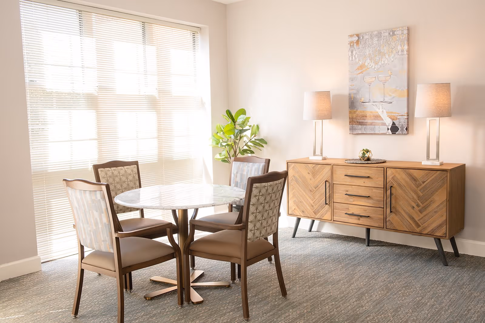 A bright dining area with a round marble table surrounded by four wooden chairs with cushioned seats and patterned backs. Behind the table is a wooden sideboard with a chevron pattern, topped with two modern table lamps and a decorative piece. A large window with blinds allows natural light to fill the room, and a green potted plant is placed in the corner.
