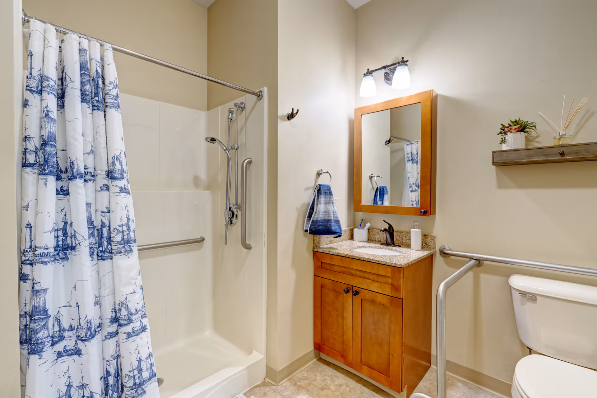A bathroom with a shower area featuring a white and blue nautical-themed shower curtain, a wooden vanity with a granite countertop, a mirror with two lights above it, a towel hanging on a ring, a toilet with a safety grab bar, and a small shelf with decorative items on the wall.