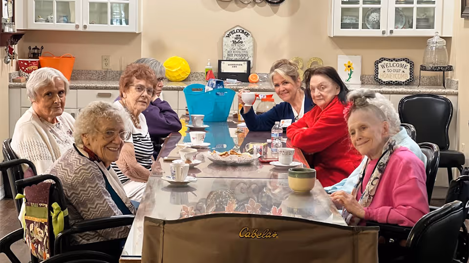 A group of elderly women sitting around a long dining table in a cozy kitchen or dining area, enjoying tea or coffee together. The table is set with cups and saucers, and there are cabinets and kitchen items in the background, including decorative signs that say 'Welcome to our Honeycomb' and 'Welcome as Hive Rules are Kind Bee Respectful Bee Positive'.
