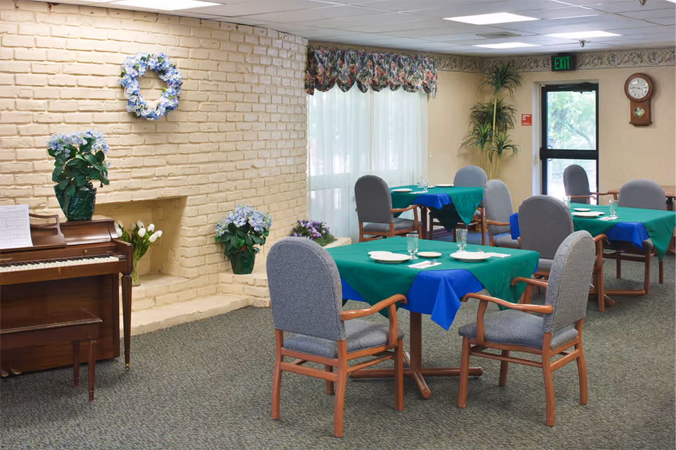 A dining room with several tables covered with green and blue tablecloths, each set with plates, glasses, and utensils. The room features a light yellow brick fireplace decorated with blue and white floral arrangements and a wreath. There is a wooden piano with sheet music on it to the left, a large window with floral valances, a clock on the wall, and a door with an exit sign above it. The chairs around the tables have wooden arms and gray upholstery.