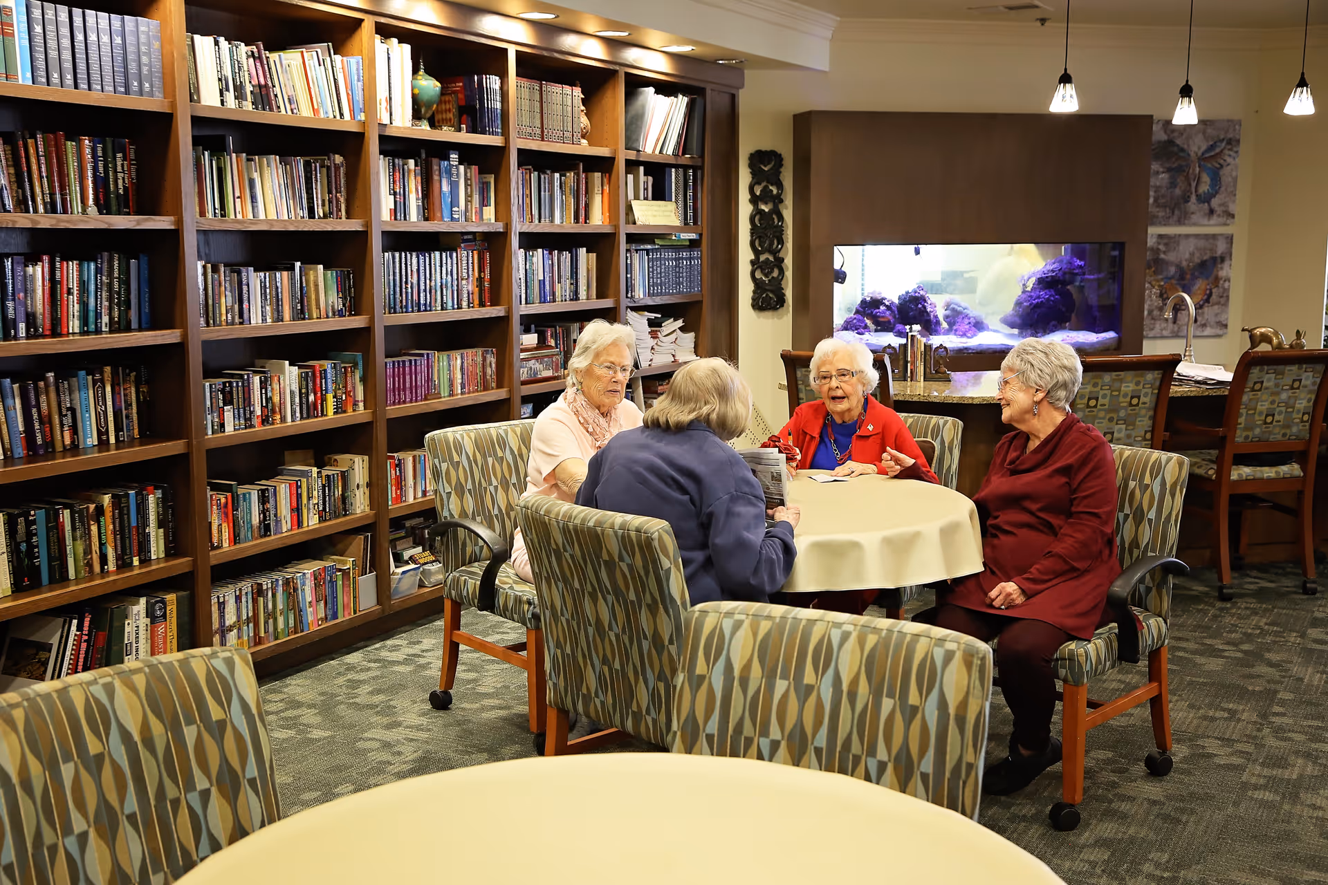 Four elderly women sitting around a round table covered with a beige tablecloth in a library or common room. Behind them are tall bookshelves filled with books, and a large aquarium with purple coral is visible in the background. The room has patterned chairs and a carpeted floor.