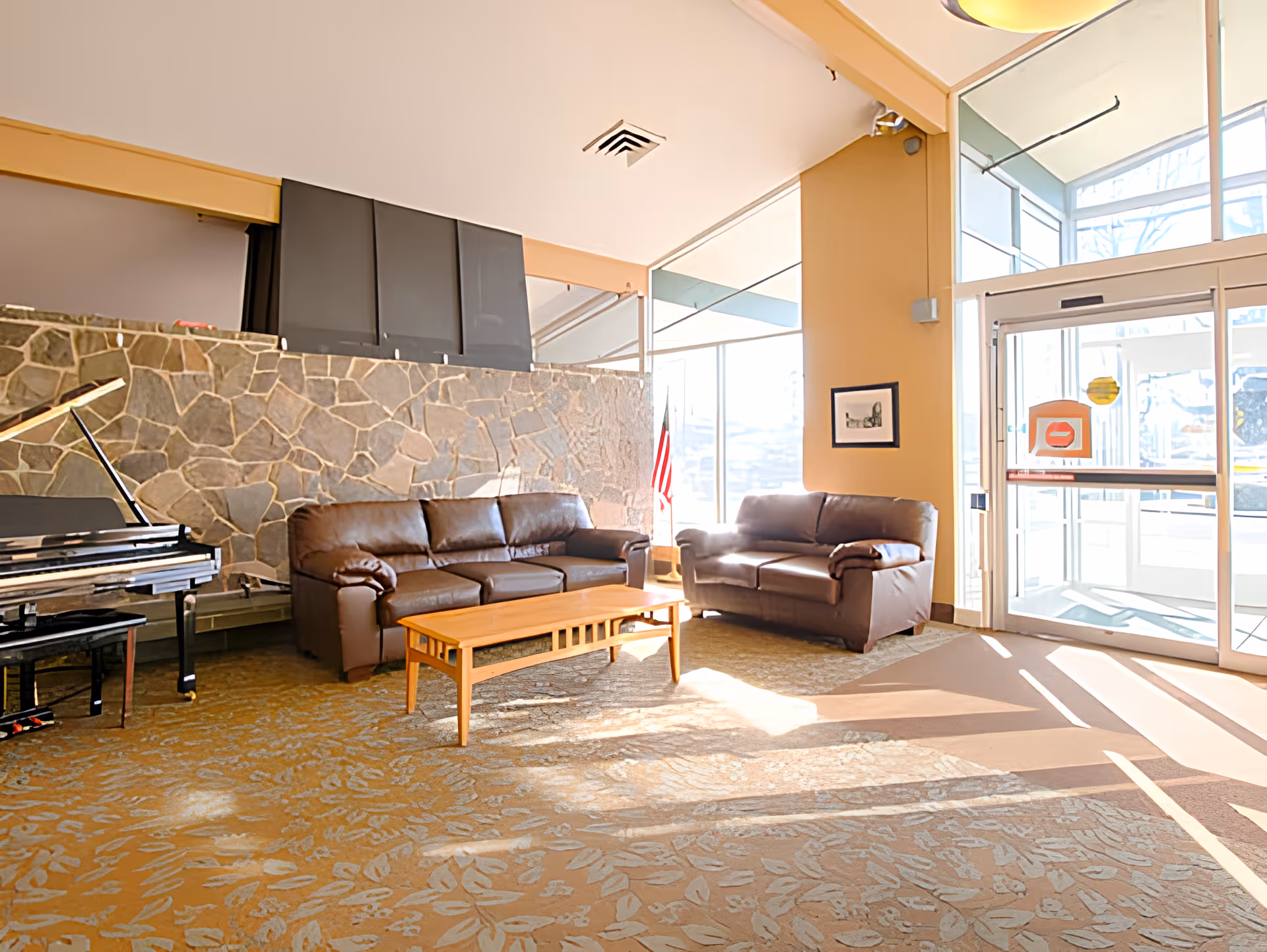 A bright and spacious lounge area with two brown leather sofas and a wooden coffee table on a patterned carpet. There is a stone wall behind the sofas and a black grand piano to the left. Large windows and glass doors allow natural light to fill the room, with an American flag visible near the windows.