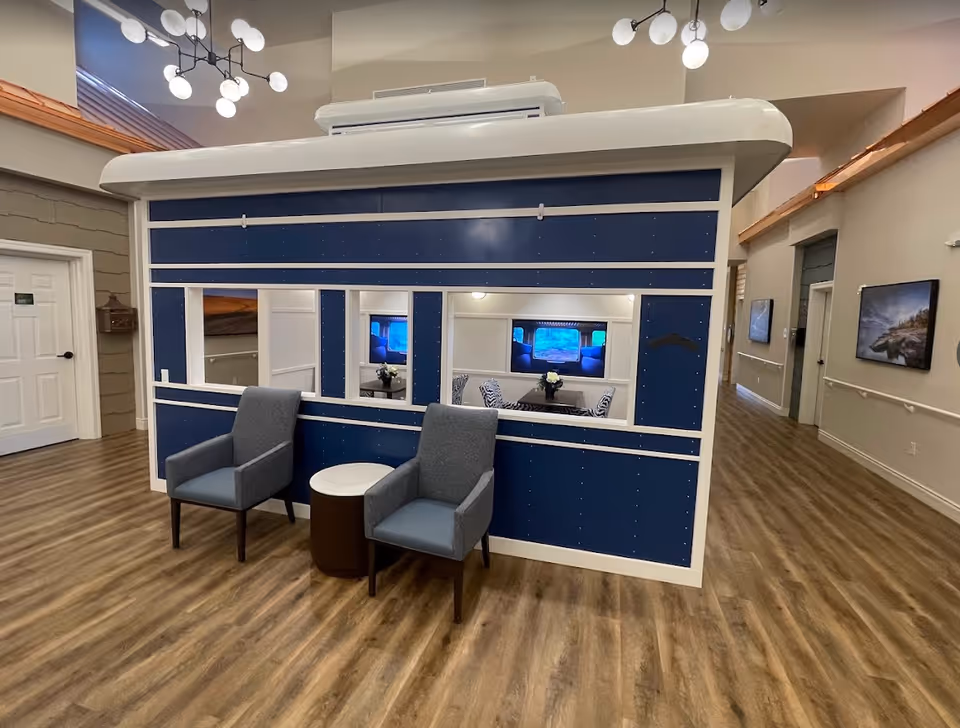 Interior common area at a memory care facility featuring a blue booth-like seating pod with windows, two gray armchairs and a small round table on wood flooring.