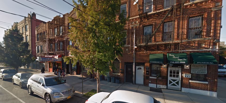 Street view of a row of brick buildings with storefronts including a barber shop and a funeral home, several parked cars along the street, and a tree on the sidewalk.